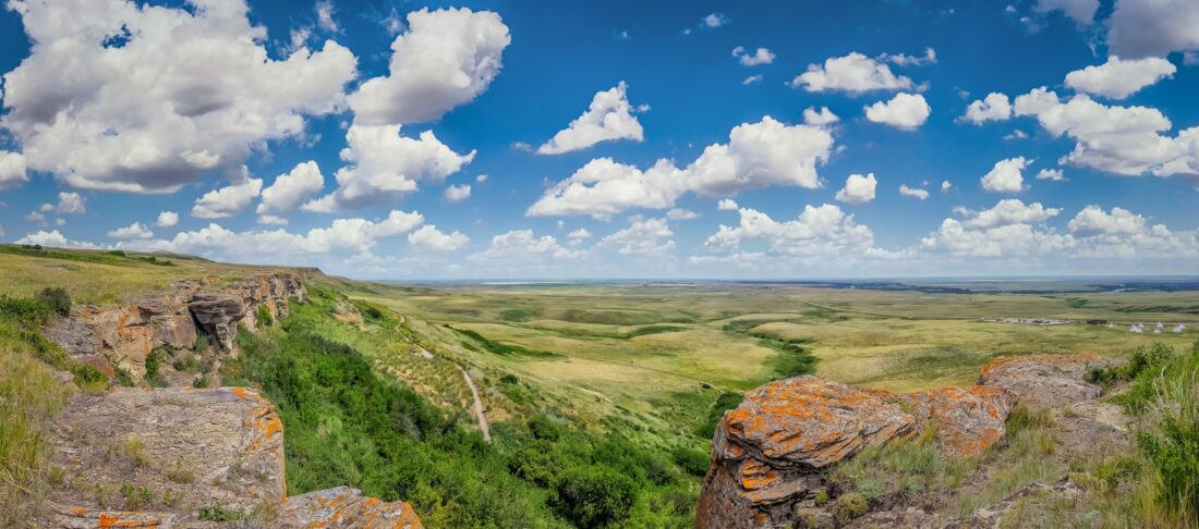 A landscape photo with green grass, blue skies, cliffs at Head Smashed in Buffalo Jump in Alberta Canada