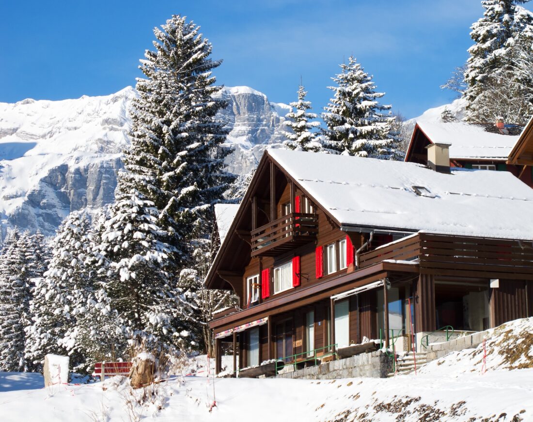 A red and brown swiss chalet house covered in snow with snow covered pine trees in the background in Switzerland.